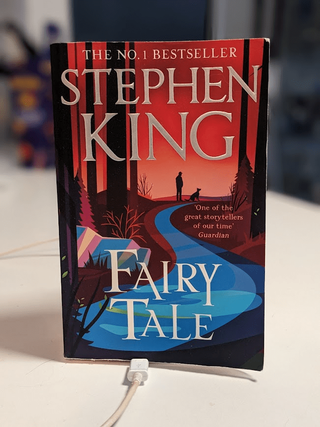A copy of the book 'Fairy Tale' (cover features a boy and a dog silhouette by a river) is standing up on a white bench.  A kindle charger appears to be plugged in to the bottom of it.