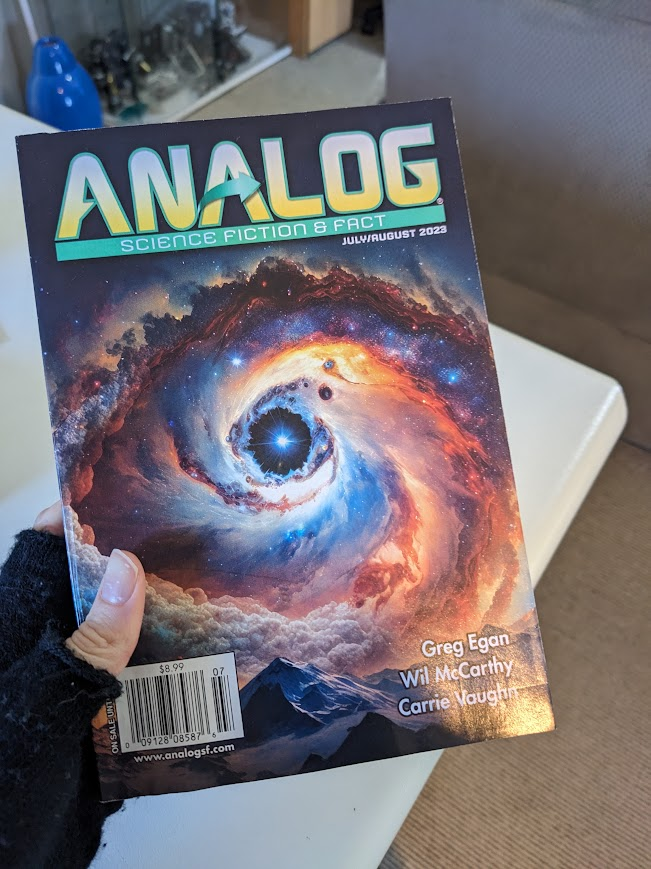 A hold holds up a paper copy of Analog Science Fiction and Fact, July/August 2023 Issue. The cover of this magazine features a swirling nebula of some sort circling around a bright star, with some mountains in the foreground.
