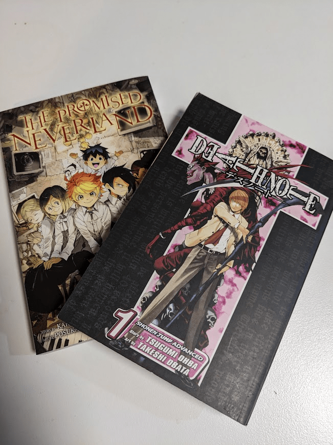 Vol. 1 of the Manga Death Note (featuring Light Yagami holding a scythe on the cover) and Vol. 7 of the Manga The Promised Neverland (Featuring the child characters sitting together) are stacked together.