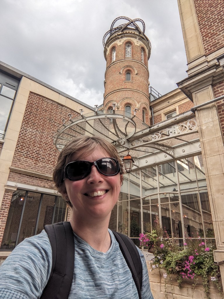 A white woman with short brown/blond hair and sunglasses takes a low-angled selfie from the courtyard in front of a large brick building. Behind her within the courtyard in a room made of glass and white iron frames. An iron circle held up by curved bars hangs over the edge of the roof of this room. In front of the glass room are bushes of pink flowers. Behind the glass room, in the interior corner of the courtyard, is a large cylindrical brick tower. Three stories of long narrow windows are visible, and on top of the tower four half-circle rings are arranged in a messy crown.