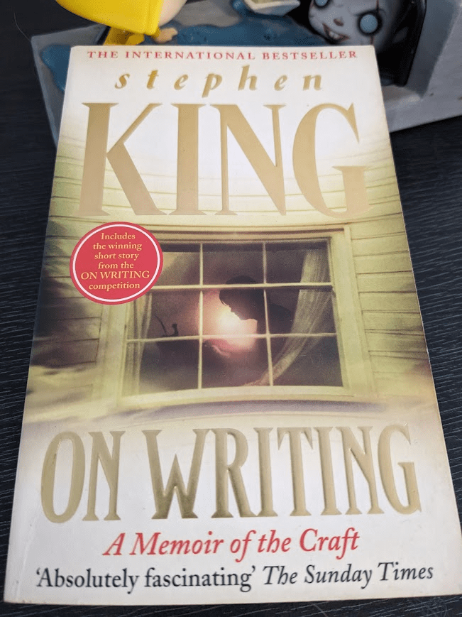 A paperback copy of Stephen King's book "On Writing, A Memoir of the Craft" leans on a Funko Pop! sculpture depicting the iconic scene of a child in a yellow raincoat crouched down in a puddle as his paper boat drifts into a gutter where a scary clown awaits.
The cover is mostly white and yellow, with the author's last name and the 'On Writing' written in large capital letters. The illustration is of Stephen King at a writing desk, seen through a glass window.