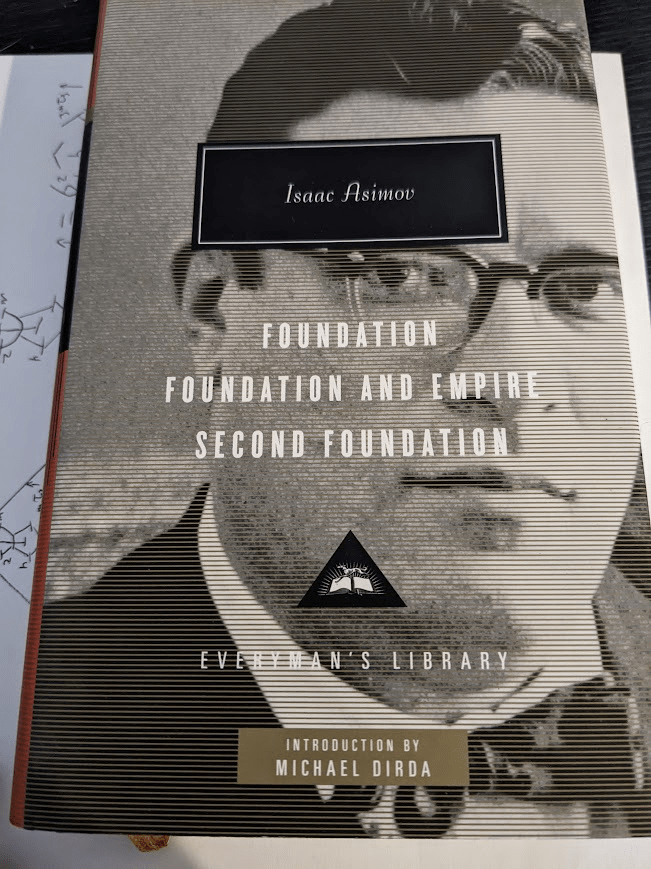A hardcover book of the Everyman's Library omnibus of Isaac Asimov's Foundation Trilogy. The cover features a close-up black and white photo of Isaac Asimov. Asimov's name is in a black box, with the titles of the trilogy (Foundation, Foundation and Empire, and Second Foundation) written beneath it. A gold box displays the words 'Introduction by Michael Dirda'. The book sits on a sheet of paper with mathematical equations written on it.