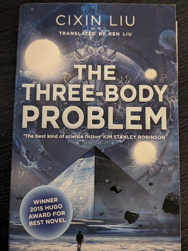 A paperback copy of "The Three-Body Problem" by Cixin Liu, translated by Ken Liu. The title is large, silver and all caps in the centre of the cover, with the following quote by Kim Stanley Robinson beneath it: "The best kind of science fiction."
The illustration beneath the author, translator and title is of a blue futuristic pyramid looming over an icy landscape surrounded by a moat. A lone human figure walks towards the pyramid, tiny in the foreground whilst massive rocks float upwards. In the background, raising into the sky and behind the title text is a strange, complex spherical device, with some orbs, and three bright suns orbiting around it.