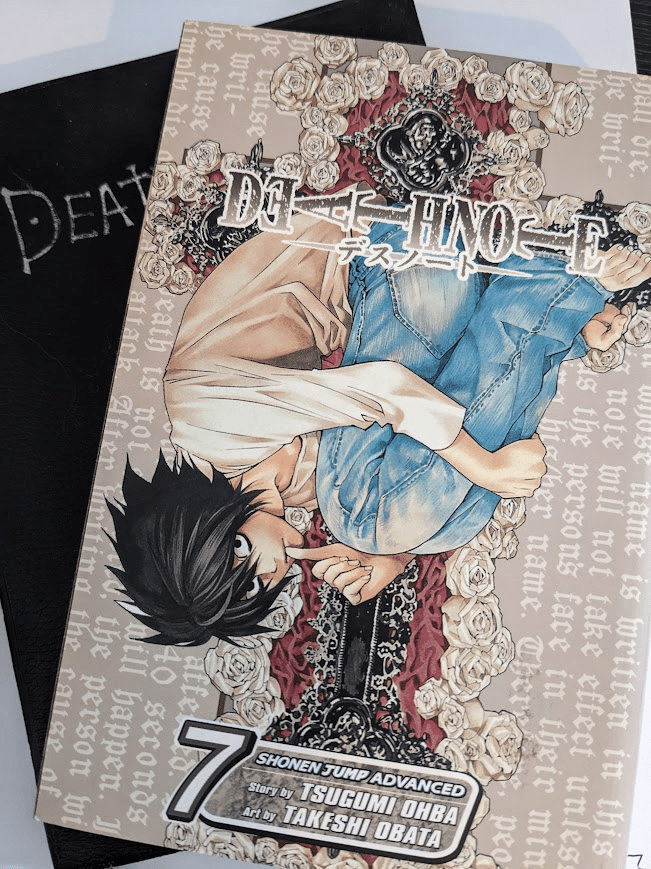 A paperback book, issue 7 of the Death Note manga, sits askew on top of a thin, leathery notebook. The heading 'DEATH NOTE' is half visible on the notebook.
The cover of Issue 7 features the character L, a black-haired young man with messy black hair wearing a white shirt and blue jeans, upside-down and curled into a fetal position with his thumb in his mouth. Behind him is an ornate gothic cross surrounded with roses on a beige background.