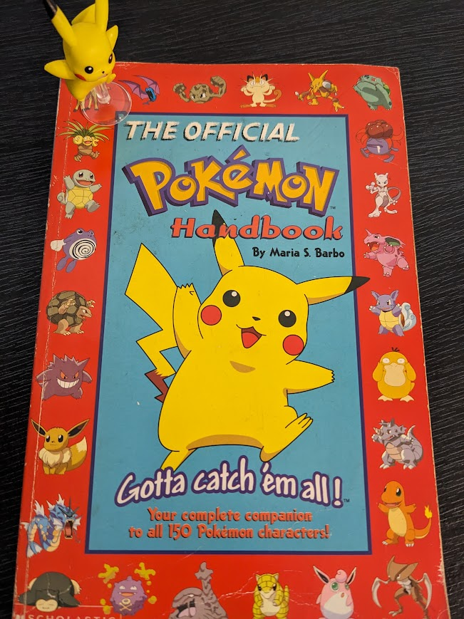 An old, battered paperback copy of "The Official Pokémon Handbook by Maria S. Barbo" sits on a black surface. The cover features a red border around a blue rectangle.
Within the rectangle the book title and authors name is at the top above a picture of Pokémon mascot Pikachu, with the tagline "Gotta catch them all - your complete companion to all 150 Pokémon characters" beneath Pikachu. About six different fonts are used for this text.
Within the red border are pictures of about two dozen different generation 1 Pokémon.
Sitting on top of the book, on the top left corner, is a small Pikachu figurine.
