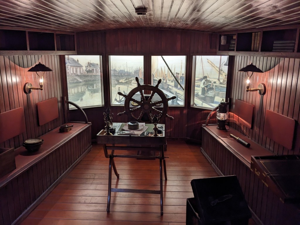 An enclosed room made entirely of dark wood that resembles a 19th Century ship's bridge. It has a ship steering wheel at the front, benches along the sides with various nautical items laid on them, and two lamps, with exposed globes and lampshades, built into the side walls. Above the lamps are overhead shelves containing books, and on the ground in the foreground is an open crate with unseen contents. At the front of the room, behind the steering wheel, where the windows/viewport should be are four paintings forming a single image of a 19th Century harbour scene. In the middle of this room is an old foldable writing desk adorned with papers and other writing paraphernalia.
