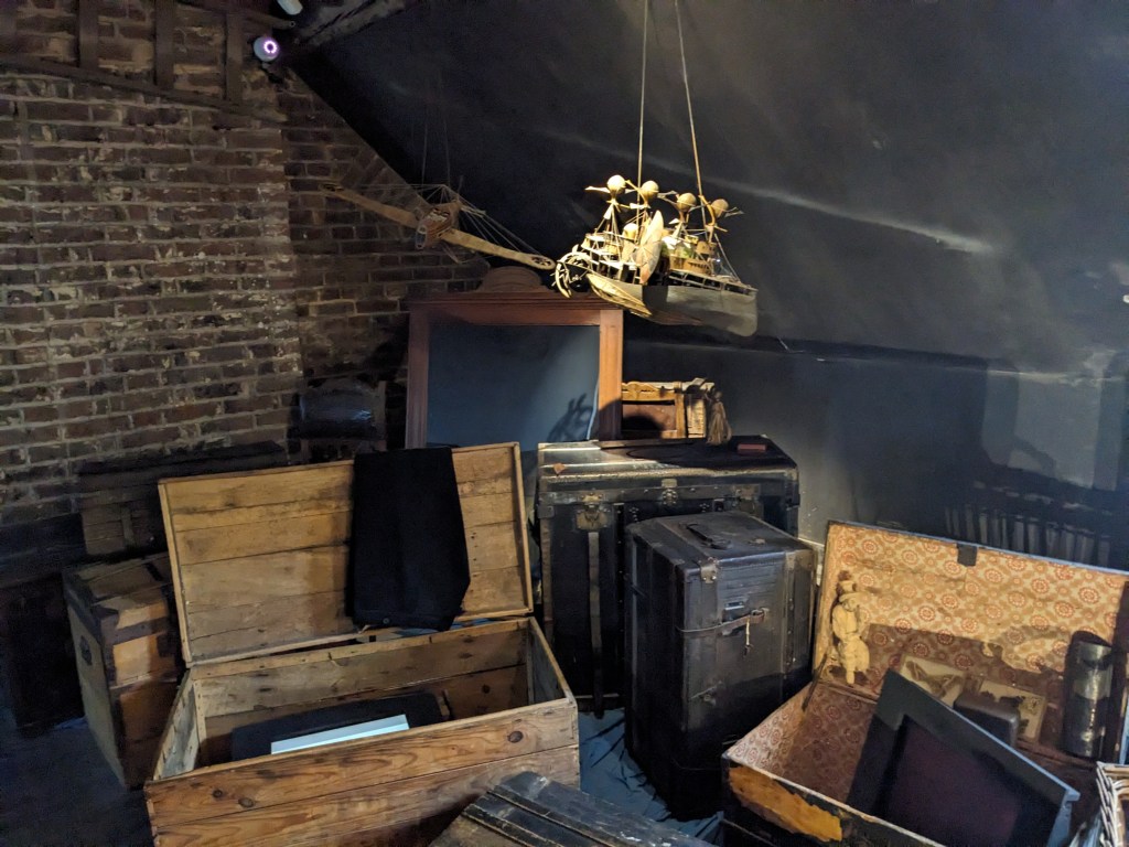 In the corner of a dark attic with brick walls, about ten 19th Century travel trunks surround a partially covered picture frame. Some of the trunks are open, displaying a range of travel related knick-knacks and souvenirs, including a doll, a thermos, some boxes, a cloak, and some preserved butterflies. Hanging from the ceiling are two models, one of a biplane, and one of a strange ship with wings, a wheel at the back, and four structures that appear to be balloons on the top.    