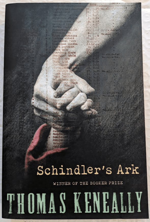 A paperback copy of 'Schindler's List' by Thomas Keneally. The illustration shows an adult hand holding the hand of a small child with a red sleeve over a grey background with a typewriter-created list of names superimposed over the image. At the bottom is the title, the words "WINNER OF THE BOOKER PRIZE" and the author's name.