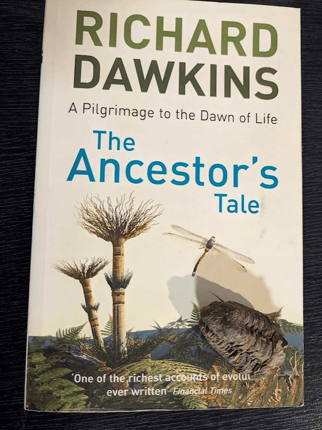 A paperback copy of 'The Ancestor's Tale' by Richard Dawkins with a small trilobite fossil placed on its lower right corner. The cover is white and has the author's name in green at the top, then the subtitle; "A Pilgrimage to the Dawn of Life" in black text, and then the title in blue. Below all the text is an illustration of a primordial scene landscape with ferns, a dragonfly, and a strange extinct plant that is comprised of a segmented trunk and wirey foliage branching outwards at the top and middle segment. At the bottom of the page, slightly obscured by the trilobite, is the following quote by the Financial Times; "One of the richest accounts of evolution ever written."