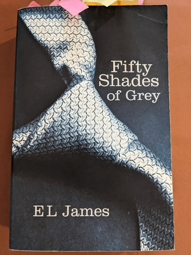 A battered paperback of 'Fifty Shades of Grey' by E.L. James. The top right corner of the book is bent, and there are dozens of red and yellow post-it notes sticking out of the top of the page. The book cover is black, with a close up photo of a silver-grey tie (mainly the know of the tie) taking up most of the page. The title is above the tie in grey text, and the author's name near the bottom of the page, also in grey.