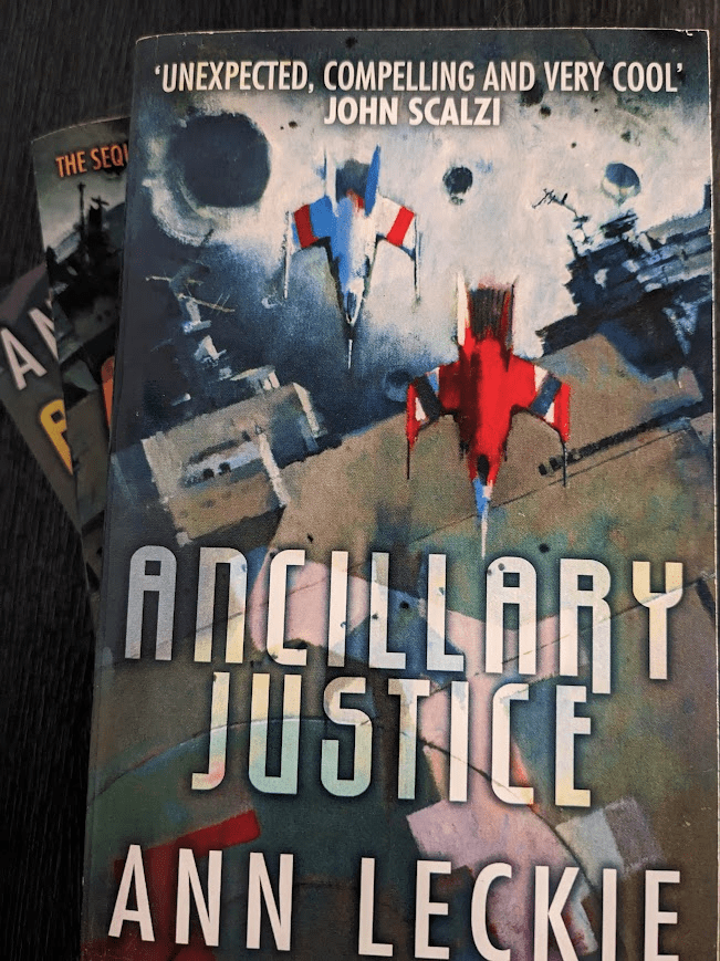 Three paperback books - the Imperial Radch Trilogy by Ann Leckie - are stacked on top of each other with 'Ancillary Justice' on top. The cover of Ancillary Justice features the title and author's name in blocky capital letters at the bottom of the page, over an illustration that shows two fighter jets (one red, one white) flying part of a massive ship with a cratered moon in the background. The shit resembles a naval battleship despite obviously being a spaceship. At the top is the following quote by John Scalzi; "Unexpected, compelling, and very cool."