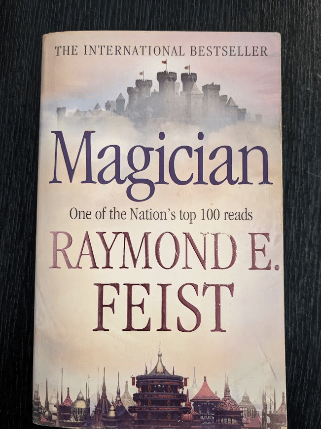 An old, slightly battered copy of 'Magician' by Raymond E. Feist. The cover illustration is pale pink (like sunset) with clouds in the centre. Over these clouds are the book title, author name the quote 'One of the Nation's top 100 reads', and the author's name. Below the clouds is an Asian inspired skyline full of stupas, domed buildings and a cylindrical tower. Above the clouds is a European style castle. The words 'The International Bestseller' are written all in caps at the top of the cover.