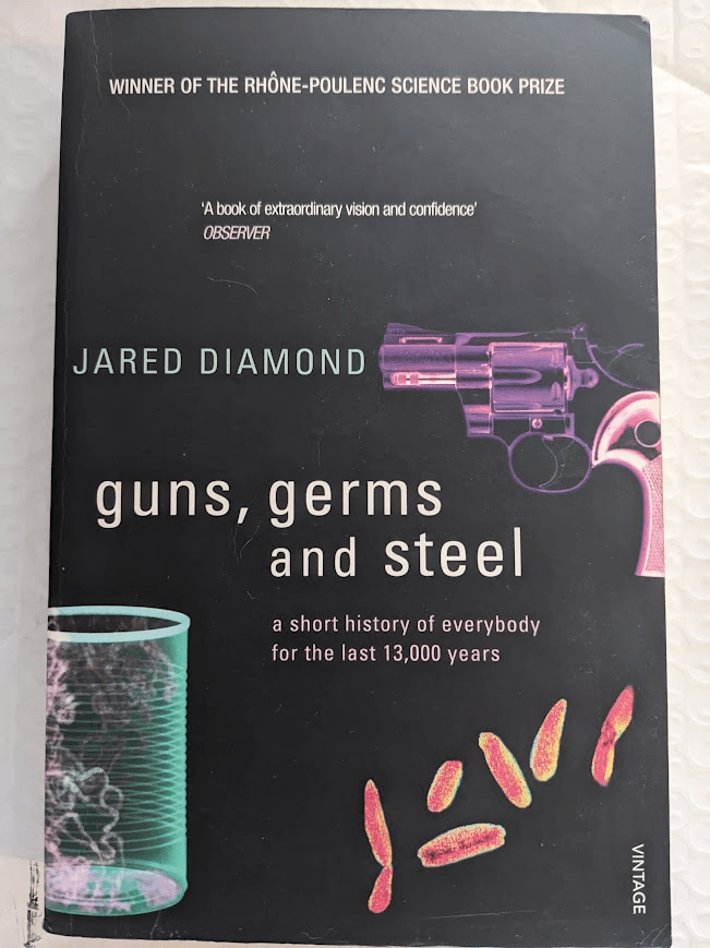 A slightly battered paperback copy of 'Guns, Germs, and Steel' by Jared Diamond. The cover is black, and images of a gun, a glass container, and some microbes are scattered around the author's name and title. At the top of the cover is the heading 'WINNER OF THE RHÔNE-POULENC SCIENCE BOOK PRIZE', with the following quote by Observer a few spaces beneath it; "A book of extraordinary vision and confidence'. Below the title is the following subtitle; "a short history of everybody for the last 13,000 years.'