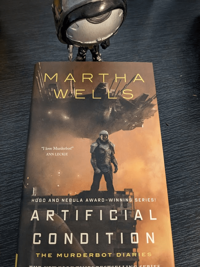 A hardcover copy of 'Artificial Condition' by Martha Wells. The cover depicts a figure in futuristic body armour including an opaque helmet standing on top of an industrial building against an orange sky. A spaceship with dozens of pointy sensors facing forward flies behind this figure. The author's name is written in yellow over the ship, while beneath the armoured figure is the title, with the subtitles 'The Murderbot Diaries' beneath it, and the heading 'HUGO AND NEBULA AWARD-WINNING SERIES' above it. The heading 'THE INTERNATIONAL BESTSELLING SERIES' is below it, but cut off. The quote 'I love Murderbot' by Ann Leckie appears halfway up the cover. Above the book stands a custom Funko Pop! figure holding a gun and resembling the Murderbot figure on the cover.