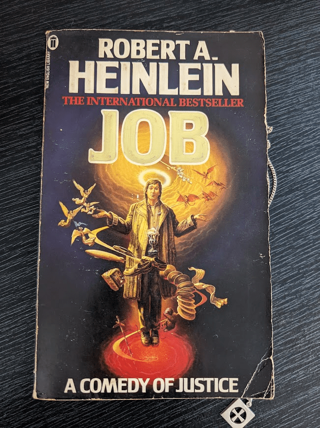 A very old, very battered paperback of 'Job: A Comedy of Justice' by Robert A. Heinlein. The bottom left corner of the cover is completely bent. The author's name is at the top of the cover, then the heading 'THE INTERNATIONAL BESTSELLER', and then the name 'JOB' in big gold letters. Beneath this heading stands a white man with brown hair, a long brown coat, and a halo, looking forward and shrugging. Swirling around him is a golden line surrounded by a chaotic mixture of items - a zeppelin, a milkshake, shoes, a bible, an ape, papers, lots of dishes, and a futuristic car driving along the line - as well as groups of angels and demons. Behind the characters head are golden heavenly clouds, while at his feet the fiery pit of hell. The subtitles "A COMEDY OF JUSTICE" is at the bottom of the cover.