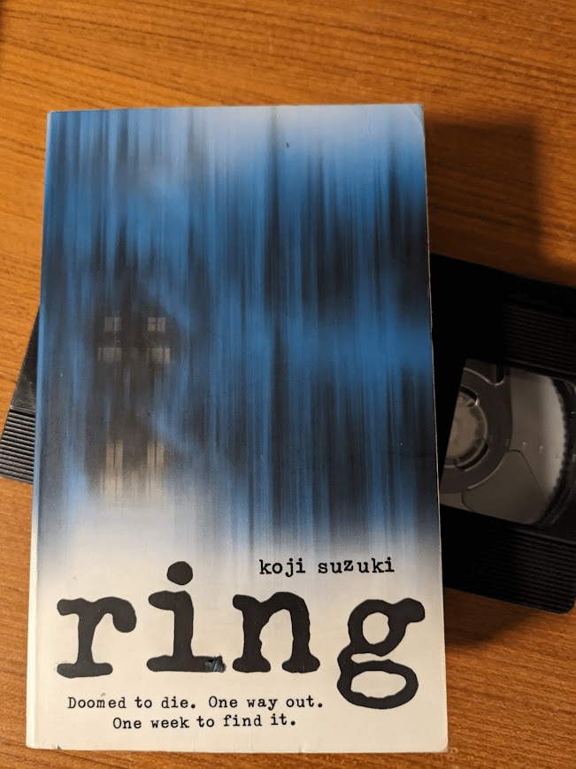 A paperback copy of 'Ring' by Koji Suzuki sits on top of a black, non-descript VHS tape. The cover is mostly blue, with streaks of black dashed vertically through it to look like rain. A tall house with lit windows looms barely visible through this 'rain'. The bottom of the cover fades to white, and has the author's name in small text, the title in big text, and the following tagline also written small; "Doomed to die. One way out. One week to find it."