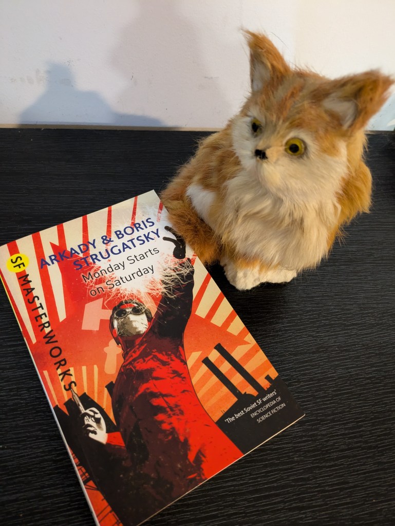 Paperback copy of the SF Masterworks edition of 'Monday Starts on Saturday' by Arkady and Boris Strugatsky. A white and orange fluffy cat figure sits next to it.