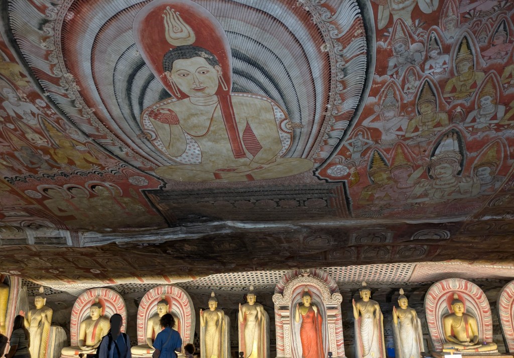 A cave with a massive, intricate fresco of Buddha siting and meditating on the ceiling, surrounded by other figures. Beneath the fresco, the walls of the cave extend further back, and a row of 10 visible Buddha statutes are lined up in an alcove. Some are sitting, some are standing.