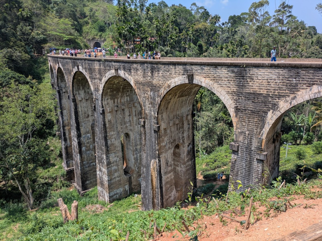 A photo of the famous nine-arch bridge near Ella, taken on a nice sunny day., Sri Lanka. The bridge is taller than the trees surrounding it, made of grey and tan bricks. Only six of the arches are visible from this angle. A large crowd of people are walking around on top of the bridge.