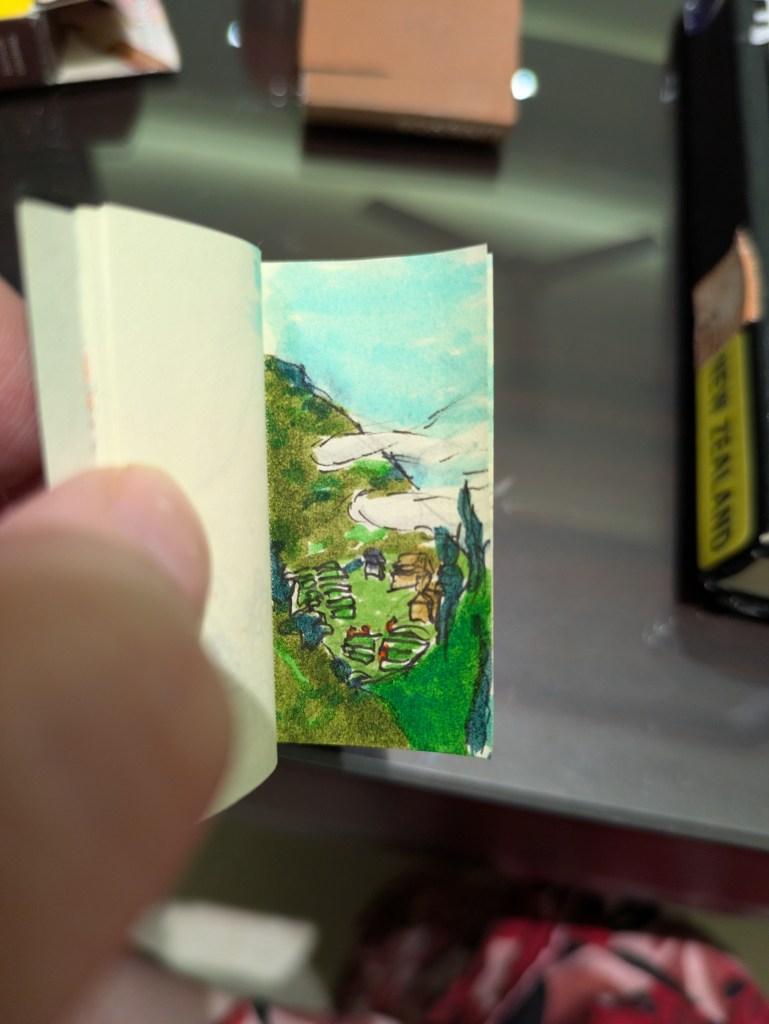 A white thumb opens a tiny book showing a drawing done in pen and marker. The drawing shows a few far away buildings and rice fields surrounded by mountains. Clouds are In front of one of the mountains.