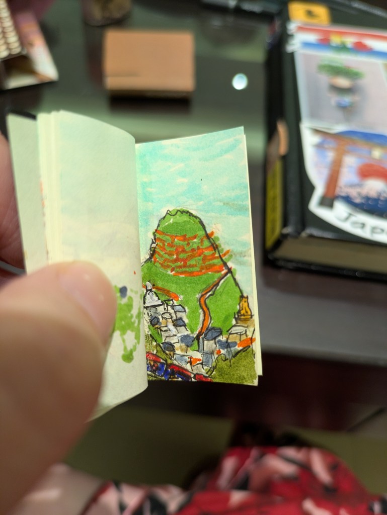 A white thumb opens a tiny book to a page with a drawing done in pen and marker. The drawing is of a green hill, with terraces for tea or rice cut into it. A path leads down the hill to a town of scattered buildings. Amongst these buildings is a Buddhist stupa and the golden top of a Hindu temple. In the foreground are two more mountains in the bottom corners, and a red and blue train going past.