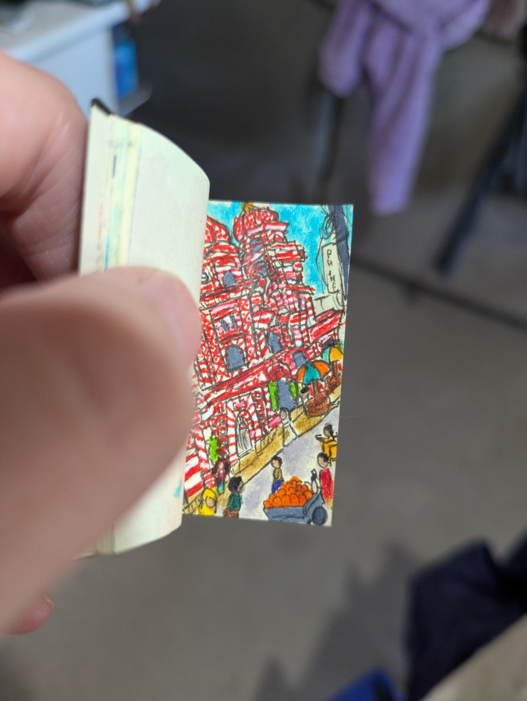 White fingers hold open a tiny book to a page with a drawing of a red and white mosque in a busy market. The mosque has two towers, and is a jumble of columns, windows, and buttresses.